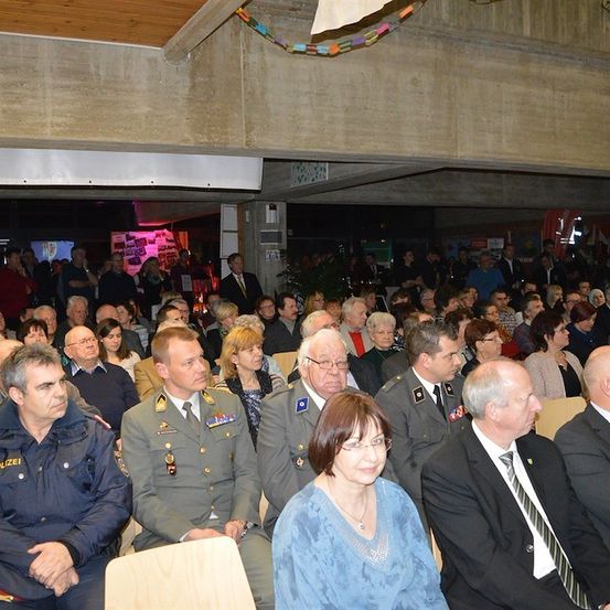 Eine große Menschenmenge, einschließlich Militärangehöriger und Zivilisten, versammelt sich in einem Saal für eine Veranstaltung. Einige sitzen, andere stehen. Der Saal hat Betonwände und eine Holzdecke.