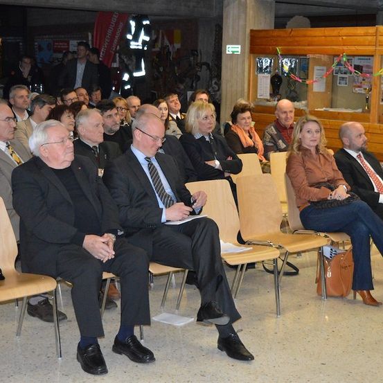 Eine Gruppe von Menschen in formeller Kleidung sitzt in einem Raum in Stühlen, möglicherweise bei einer Versammlung oder Veranstaltung. Einige schauen auf Papiere, während andere in Gespräche vertieft sind.