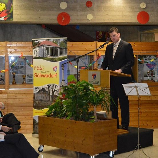 Ein Mann in Anzug und Krawatte steht an einem Rednerpult mit Mikrofon und hält eine Rede. Hinter ihm befindet sich eine Holzkulisse mit einem Banner und einem Topf mit Pflanzen. Im Hintergrund sitzen Menschen, und eine Bühne mit einem Notenständer ist sichtbar.