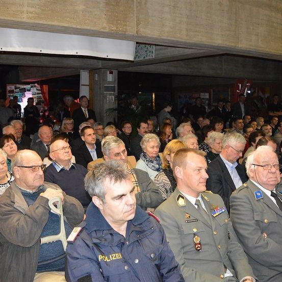 Eine große Gruppe von Menschen, darunter Männer und Frauen, sitzt in einem Saal. Einige tragen Brillen und einige tragen Militäruniformen. Sie nehmen wahrscheinlich an einer formellen Veranstaltung teil.