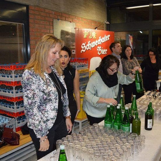 Eine Gruppe von Menschen versammelt sich um einen Tisch voller Flaschen und Gläser, möglicherweise in Vorbereitung auf eine Veranstaltung. Eine Frau in einer Blumenjacke steht neben einer Frau mit Brille, die eine Flasche hält. Hinter ihnen steht ein Schild mit der Aufschrift 'Schwedwetter Bier'.
