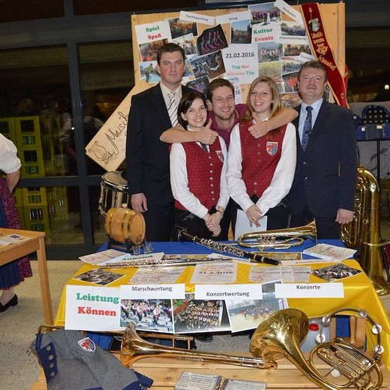 Eine Gruppe von Menschen steht um einen Tisch mit Musikinstrumenten und einem Schild, auf dem 'Leistung Können' steht. Sie sind in passenden Westen gekleidet und scheinen für ein Foto zu posieren. Dahinter befindet sich eine Pinnwand mit Bildern und Text.