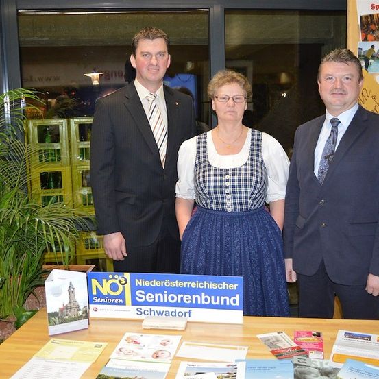 Drei Personen stehen an einem Tisch mit Werbematerialien, darunter ein Schild, das 'Niederosterreichischer Seniorenbund Schwadorf' besagt. Der Mann links trägt einen Anzug und eine Krawatte, die Frau in der Mitte trägt eine Brille und eine Halskette, und der Mann rechts trägt einen Anzug und eine Krawatte. Hinter ihnen ist eine Glastür mit einer Reflexion eines Gebäudes.