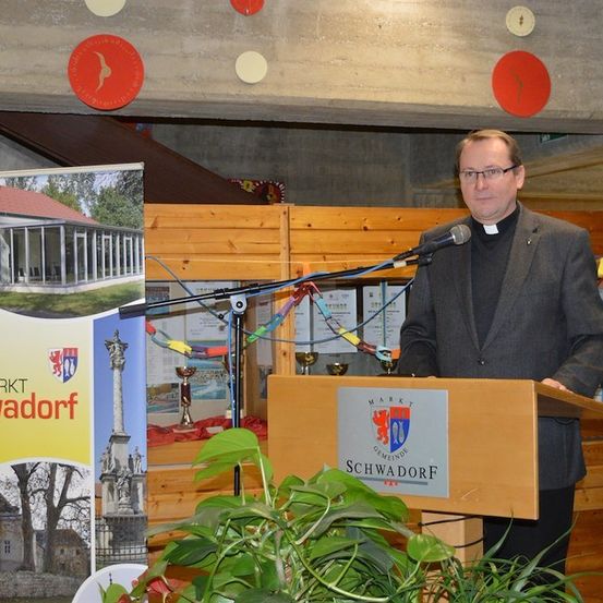 Ein Priester steht an einem Podium mit einem Mikrofon in einer Kirche, mit einem Schild für Markt Schwadorf im Hintergrund.