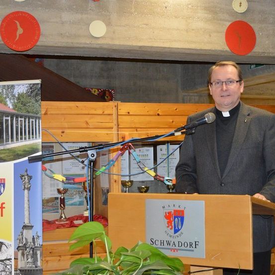 Ein Mann steht an einem Rednerpult mit Mikrofon, trägt einen Klerikerkragen und scheint eine Rede zu halten. Hinter ihm befindet sich ein hölzerner Hintergrund mit einem Schild, auf dem 'Marktgemeinde Schwadorf' steht. Links sind eine Pflanze und ein Plakat zu sehen.
