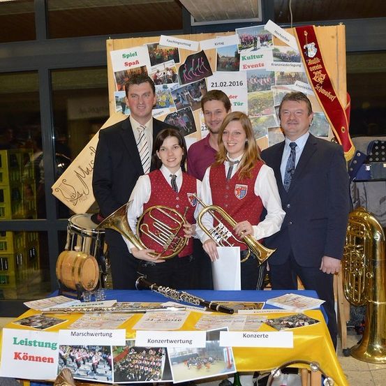 Eine Gruppe von Musikern, gekleidet in traditioneller Kleidung, steht hinter einem Tisch mit Musikinstrumenten und Werbematerialien. Der Tisch hat Schilder, die 'Leistung Können' und 'Konzerte' lesen. Dahinter ist eine Wand mit verschiedenen Fotos und einem Banner.