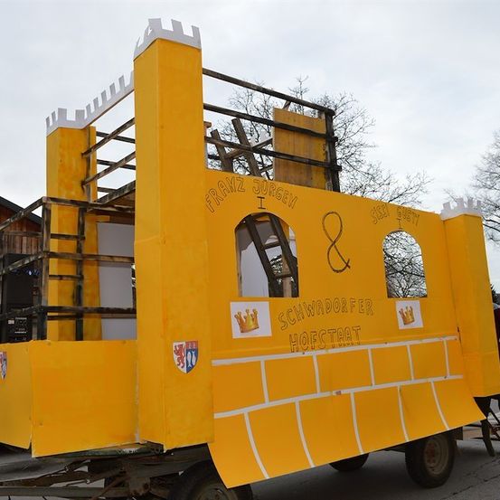 Ein gelber Wagen mit einer burgartigen Struktur auf einem Anhänger, mit den Texten 'Franz Jurgen I' und 'Schwarzdorfer Hofstaat'. Links befindet sich ein Lautsprecher, rechts steht eine Person.