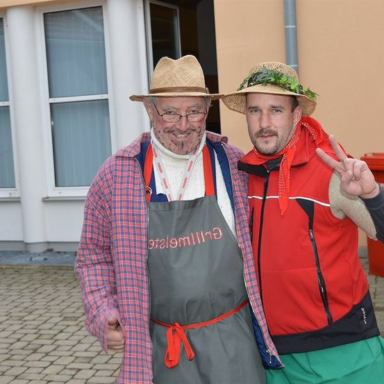 Zwei Männer in Kostümen, einer mit einer Kochschürze und der andere in einer roten Jacke, stehen vor einem Gebäude.