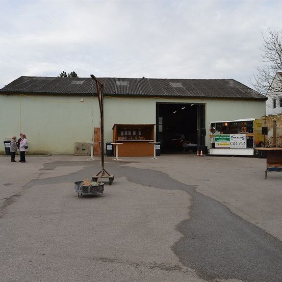 Ein Außenbereich mit einem Gebäude, einem Imbisswagen und einer kleinen Struktur mit einem Feuerkorb davor. Zwei Personen stehen in der Nähe des Gebäudes.