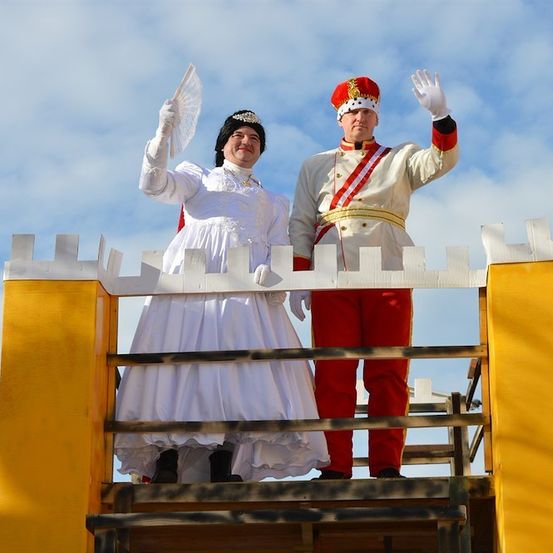 Ein Mann und eine Frau in historischen Kostümen stehen auf einer Bühne und winken der Menge zu. Die Frau hält einen weißen Fächer.