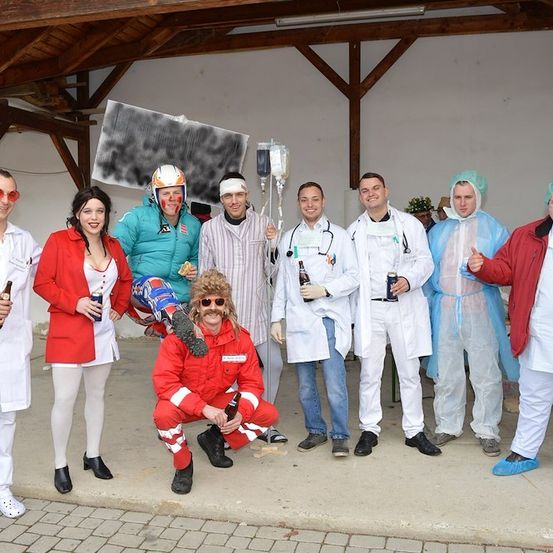 Eine Gruppe von Menschen in verschiedenen Kostümen, einschließlich medizinischer Kleidung, posiert für ein Foto in einer Scheune. Einige halten Bierflaschen und eine Infusionsflasche.