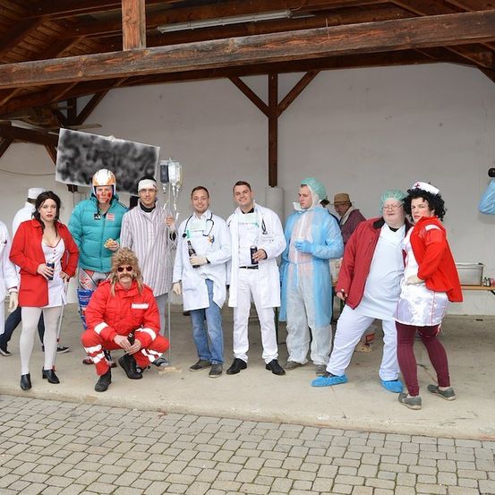 Eine Gruppe von Menschen in verschiedenen Kostümen, darunter Laborkittel, Feuerwehrmann-Anzüge und Clownskostüme, posiert für ein Foto in einem überdachten Außenbereich. Einige halten Flaschen und tragen Masken.