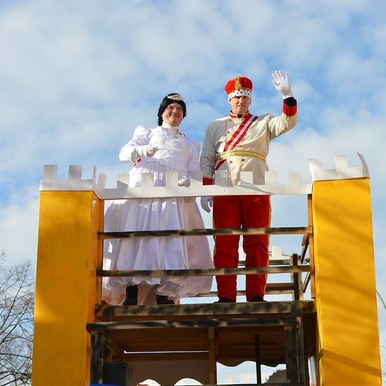 Ein Mann und eine Frau in königlicher Kleidung stehen auf einer gelben Plattform und winken der Menge zu. Der Mann trägt ein rot-weißes Uniform, während die Frau ein weißes Kleid trägt. Sie stehen auf einem Wagen, wahrscheinlich für eine Parade.