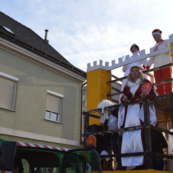 Drei Personen in mittelalterlichen Kostümen stehen auf einem geschmückten Wagen vor einem Gebäude.