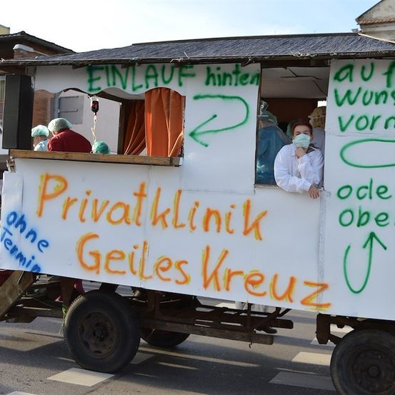 Ein dekorierter Wagen mit 'Privatklinik Geiles Kreuz' darauf, mit Personen in medizinischen Gewändern im Inneren und Pfeilen, die auf verschiedene Richtungen an der Seite zeigen.