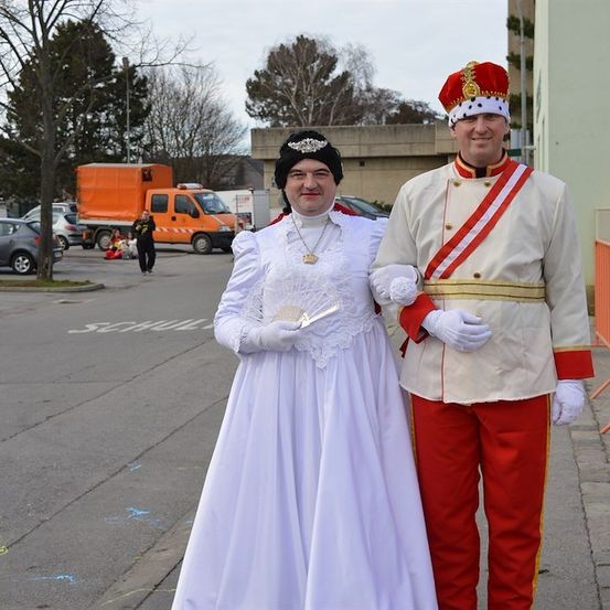 Ein Mann und eine Frau in historischen Kostümen stehen zusammen auf einer Straße. Der Mann trägt ein rot-weißes Uniform mit Krone, und die Frau trägt ein weißes Kleid mit Krone und hält einen Fächer. Im Hintergrund sind verschwommen geparkte Autos, ein Lastwagen und Bäume zu sehen.