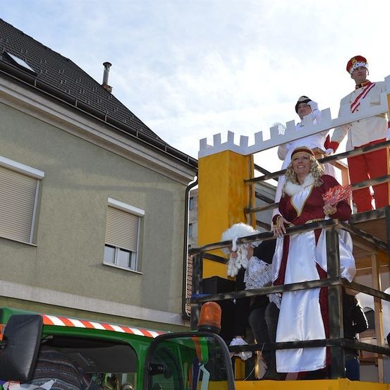 Drei Personen in mittelalterlichen Kostümen stehen auf einem Festwagen vor einem Gebäude, eine hält einen Blumenstrauß.