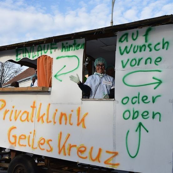 Eine Person in einer medizinischen Uniform steht in einer Kabine mit Graffiti an den Wänden. Die Graffiti enthalten Wegbeschreibungen und erwähnen 'Privatklinik Geiles Kreuz.' Die Person trägt einen Haarnetz und Handschuhe.