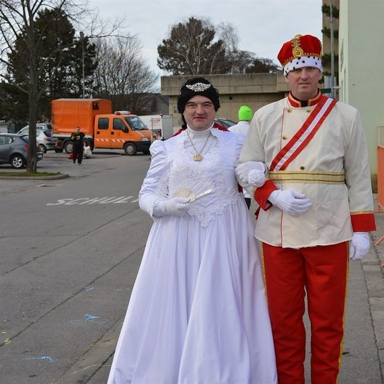 Ein Mann und eine Frau in königlichen Kostümen stehen auf einer Straße, mit einem verschwommenen Hintergrund von geparkten Autos und einem Gebäude.