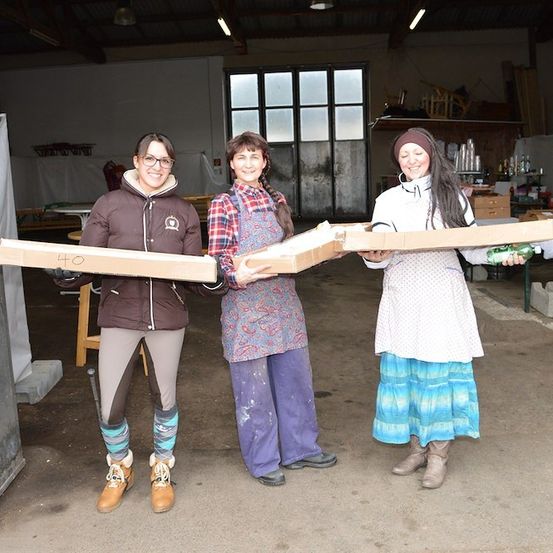 Drei Frauen in einer Werkstatt, jede hält eine große Pappschachtel und posiert für ein Foto. Die Frau links trägt eine Brille und eine Jacke, die mittlere ein kariertes Hemd und eine Schürze und die rechte ein weißes Kleid.