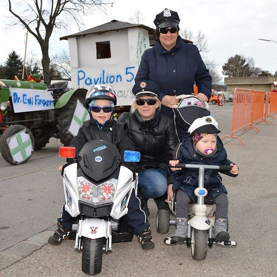 Eine Familie in Polizeiuniformen, mit zwei Kindern auf kleinen Fahrrädern und einer Frau, die ein Baby in einem Kinderwagen hält. Dahinter steht ein Traktor und ein Gebäude mit einem Schild, das Pavillon 5 liest.