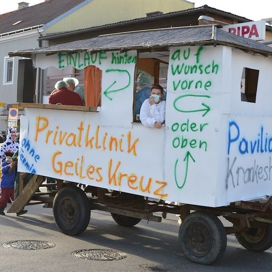 Ein weißer Anhänger mit Graffiti, auf dem 'Privatklinik' und 'Geiles Kreuz' steht, wird von einem Pferd gezogen. Eine Person mit Maske ist drinnen.