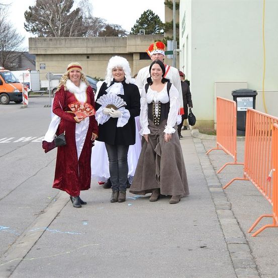 Vier Menschen in historischen Kostümen stehen auf einem Bürgersteig und lächeln für ein Foto. Hinter ihnen befinden sich orangefarbene Barrieren und ein Mülleimer. Im Hintergrund steht ein Gebäude, ein Lieferwagen und Bäume.