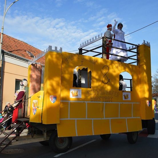 Ein gelber Umzugswagen mit mittelalterlichem Thema steht auf der Straße, mit Menschen in Kostümen, die winken. Der Wagen hat eine burgartige Struktur oben mit einem Paar, das winkt. Der Wagen ist mit mittelalterlichen Symbolen und Text verziert.