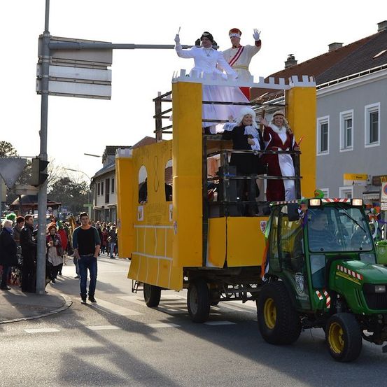 Ein farbenfroher Wagen mit kostümierten Personen wird von einem grünen Traktor in einer Parade gezogen. Zuschauer säumen die Straße.