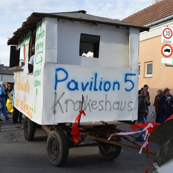 Ein dekorierter Anhänger mit der Aufschrift 'Pavillon 5 Krakeshaus', um den herum Menschen stehen. Der Anhänger ist weiß gestrichen und mit blauer und grüner Graffiti verziert. Eine Person ist im Anhänger.