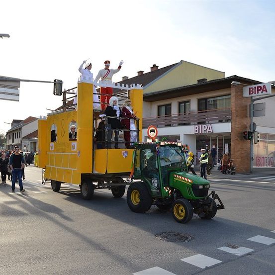 Ein Paradewagen mit kostümierten Personen wird von einem grünen Traktor auf einer Straße gezogen. Der Wagen ist gelb und weiß, und es gibt Gebäude und Menschen, die auf dem Bürgersteig gehen.