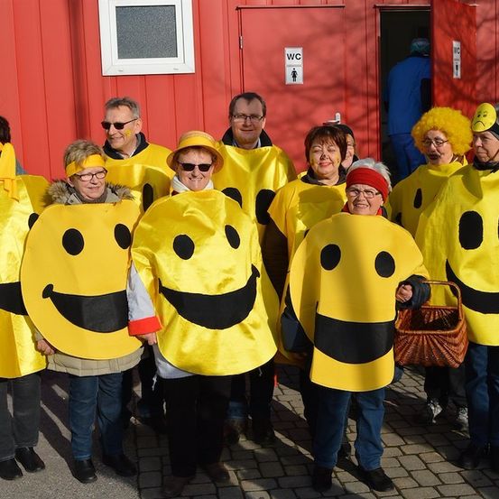 Eine Gruppe von Erwachsenen in gelben Smiley-Kostümen steht vor einem roten Gebäude.