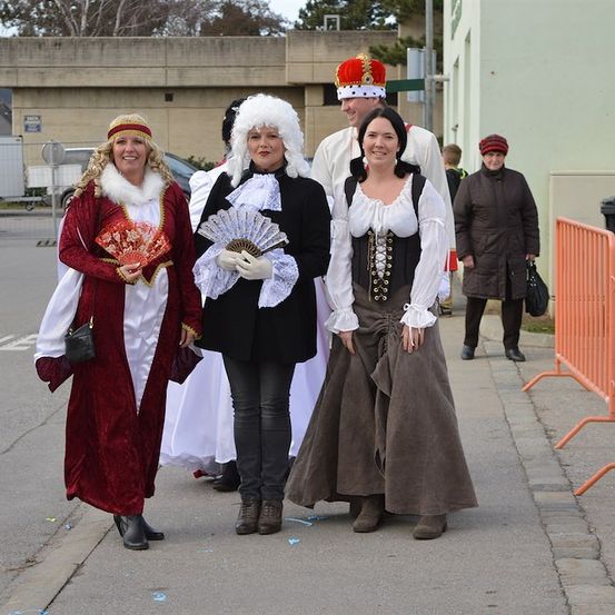 Drei Frauen in historischen Kostümen gehen eine Straße entlang, ein Mann mit Krone hinter ihnen. Eine Frau in einer braunen Jacke steht neben einem Mülleimer.