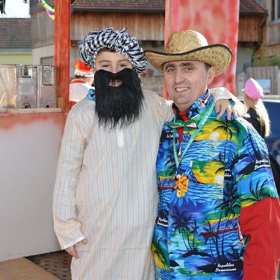 Zwei Männer in Kostümen, einer mit Turban und künstlichem Bart, der andere in einem Hawaiihemd, stehen lächelnd für ein Foto.