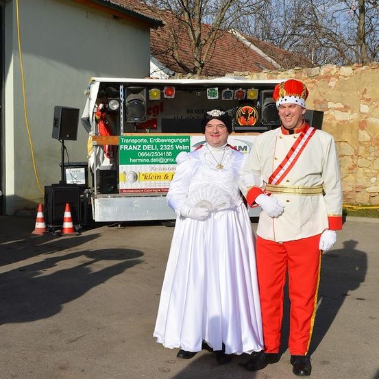 Zwei Personen in Kostümen, eine in einem weißen Kleid und die andere in einer roten und weißen Uniform, stehen vor einem Fahrzeug mit einem Schild, das 'Transporte, Erdbwegungen Franz Dell, 2325 Velm' besagt. Dahinter befindet sich eine Steinmauer und ein Haus mit einem Ziegeldach.