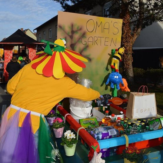 Eine Person in einem bunten Kostüm steht hinter einem Wagen mit Blumen, Flaschen und einem Schild, das Oma's Garten sagt.
