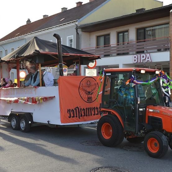 Ein Traktor mit einem orangefarbenen Banner zieht einen Anhänger mit einer Markise und Leuten darauf. Der Traktor steht vor einem Gebäude mit dem Schild BIPA.