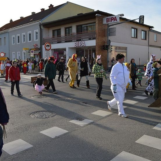 Eine Straße voller Menschen in Kostümen, einige gehen über einen Zebrastreifen, mit Gebäuden und einem Schild für BIPA im Hintergrund.