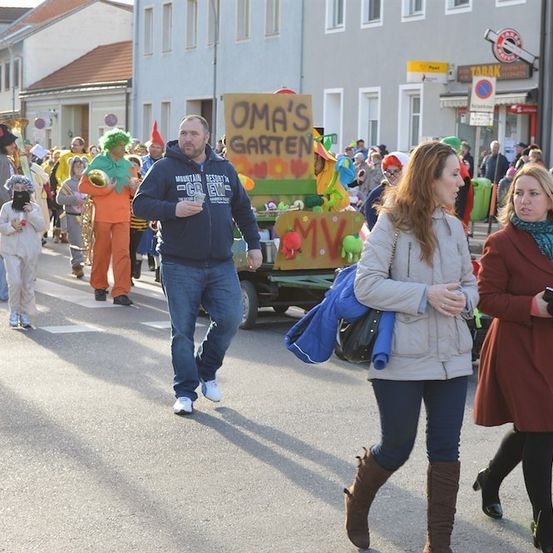 Ein Straßenumzug mit Menschen in Kostümen, die in der Mitte der Straße gehen, einem Mann in einer blauen Kapuzenjacke und zwei Frauen in Mänteln, die auf der rechten Seite etwas betrachten.