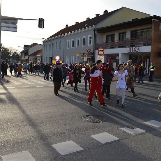 Eine Gruppe von Menschen in Kostümen geht über eine Straße, mit Gebäuden und einer Ampel im Hintergrund.
