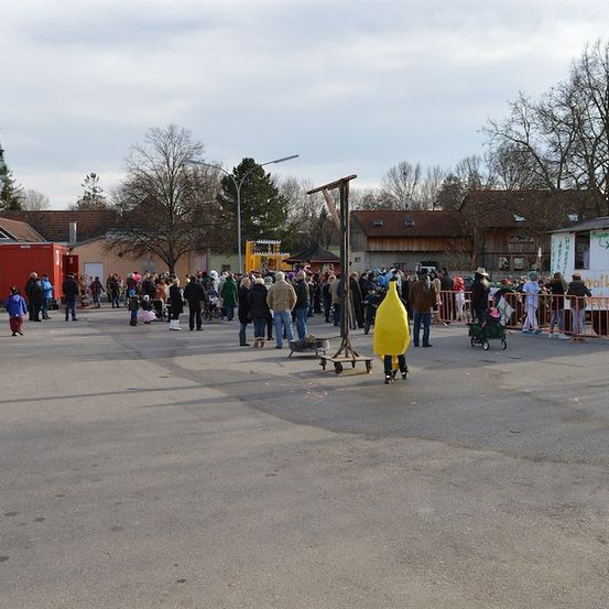 Eine Menschenmenge versammelt sich in einem Außenbereich, wahrscheinlich für eine Veranstaltung. Einige tragen Winterkleidung, andere Kostüme. Eine Person in einer gelben Bananen-Kostümierung steht in der Nähe eines Zauns. Im Hintergrund befinden sich mehrere Gebäude und Bäume.