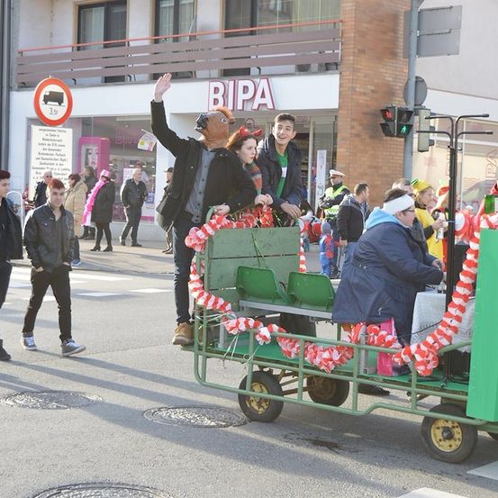 Eine Gruppe von Menschen in Kostümen und Masken fährt in einem geschmückten grünen Wagen bei einer Parade auf einer Straße.