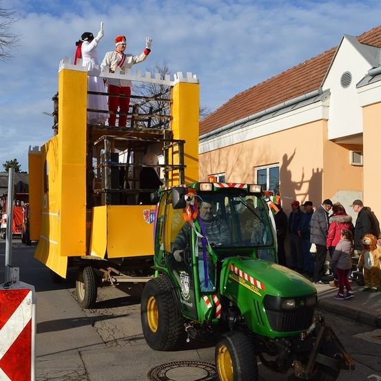 Ein grüner Traktor mit einem gelben Anhänger fährt eine Straße hinunter, an der Menschen auf dem Bürgersteig stehen. Auf dem Anhänger stehen zwei Menschen auf einer Plattform in Kostümen.