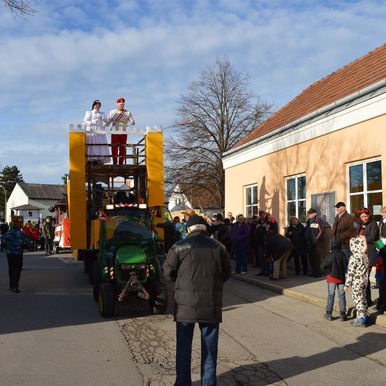 Ein Umzug mit einem geschmückten Traktor, auf dem zwei Personen in Kostümen sitzen. Eine Menschenmenge beobachtet am Straßenrand.