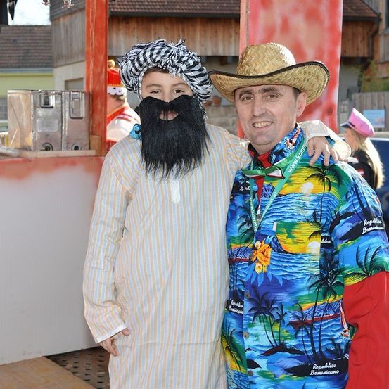 Zwei Männer in Kostümen, einer in einem Turban und mit einer falschen Bart, der andere in einem tropischen Hemd, stehen zusammen vor einem hölzernen Stand.