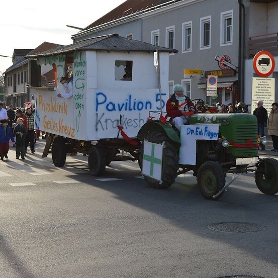 Ein Traktor, der für einen Umzug dekoriert ist, mit 'Pavillon 5' und 'Dr. Edi Finger' darauf geschrieben, mit Leuten in Laborkittel, die dahinter gehen.