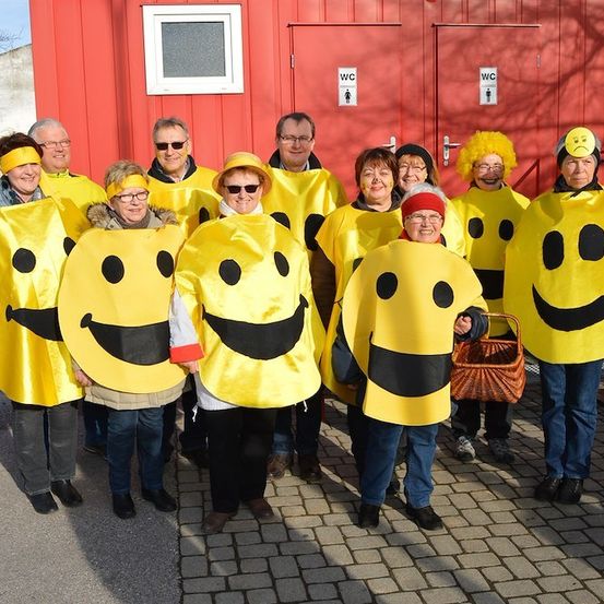 Eine Gruppe von Menschen in gelben Smiley-Kostümen steht vor einem roten Gebäude mit WC-Schildern an den Türen.