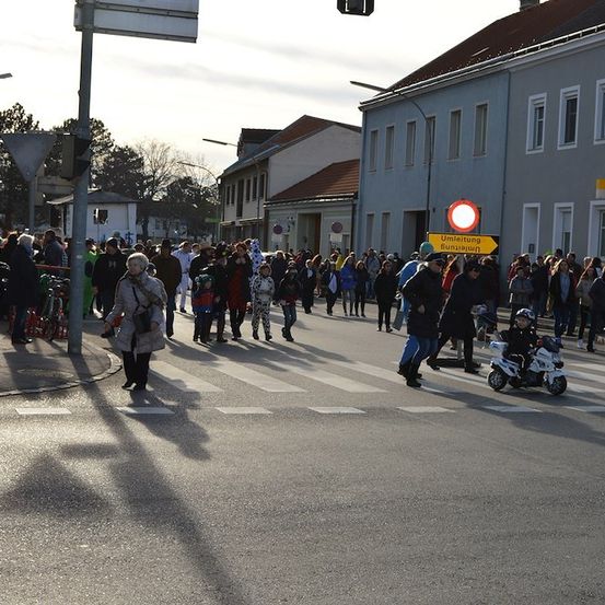 Eine große Menschenmenge geht über eine Straße, einige tragen Kostüme. Ein Kind fährt einen Roller.