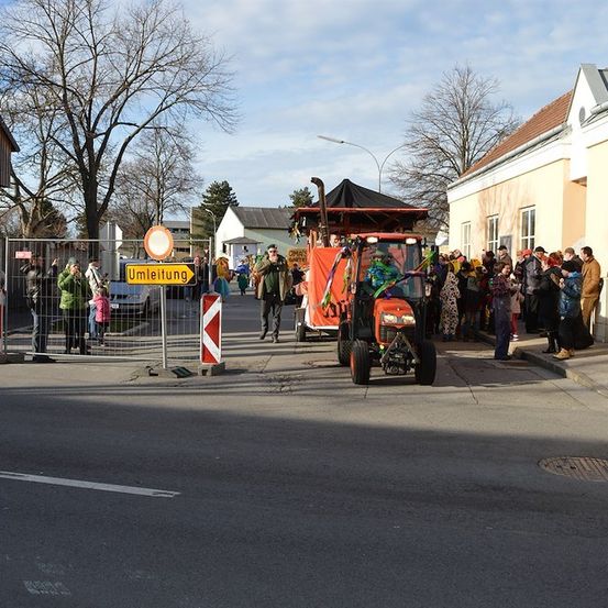 Ein roter Traktor mit einem Baldachin fährt durch einen Stadtplatz, mit Menschen auf dem Bürgersteig und einem Straßenschild, das 'Umleitung' bedeutet.