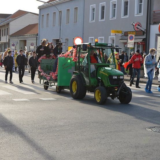 Ein grüner Traktor, der mit Blumen und Bändern verziert ist, fährt mit Menschen hinter ihm her. Der Traktor zieht einen Anhänger, auf dem Menschen sitzen. Die Menschen tragen Kostüme. Es gibt Gebäude an der Straßenseite.